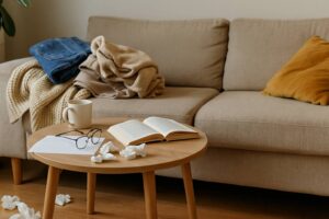 Beigefarbenes Sofa mit Decke und Jeans, davor ein kleiner Holztisch mit Tasse, Brille und offenem Buch – eine ruhige Alltagsatmosphäre, die zeigt, wie Unordnung und Gemütlichkeit zusammenwirken.